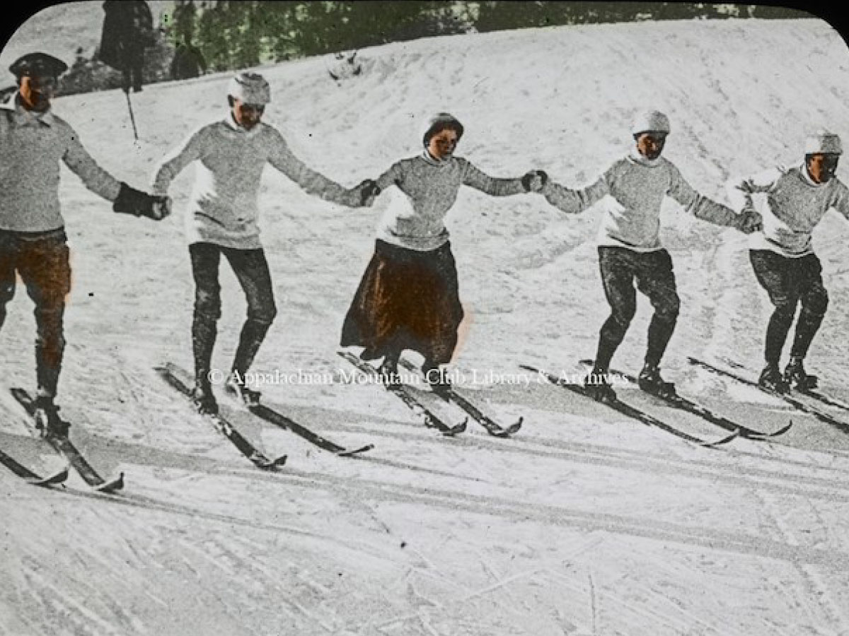 Five skiers in a line, holding hands and descending a hill